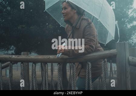 Frau lächelt und genießt es, an einem regnerischen Tag unter dem Regenschirm im Freien zu parken. Trendige Frau in der Wintersaison in der Freizeit Aktivität unter dem Regen. Menschen und schlechtes Wetter im Freien. Stockfoto