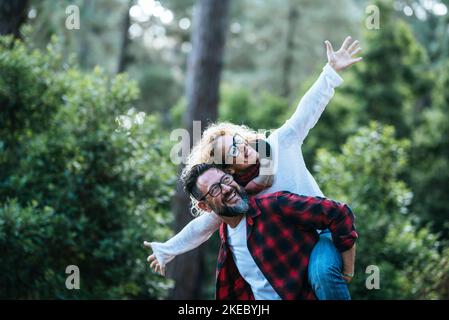 Überglücklich und glücklich Paar haben Spaß zusammen in der Park Natur. Mann tragen Frau im Huckepack in Outdoor-Freizeitaktivitäten. Grüner Wald im Hintergrund. Hipster Person lächeln und lachen viel im Freien Stockfoto
