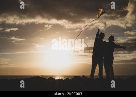 Nahaufnahme und Porträt von zwei alten und reifen Menschen, die am Strand mit dem Meer im Hintergrund und Sonnenuntergang mit einem flaying Kite spielen und genießen - aktive Senioren, die Spaß in der Silhouette haben Stockfoto