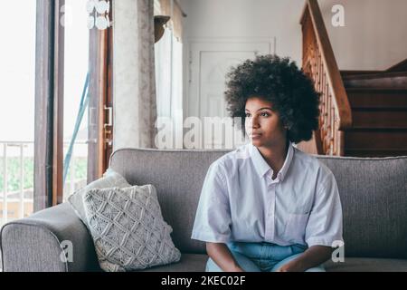 Nachdenkliche afroamerikanische Frau, die auf einem bequemen Sofa sitzt und durch das Fenster nach draußen blickt. Ernsthafte nachdenkliche schwarze Dame, die sich auf der Couch entspannt und die Aussicht vom Hausfenster bewundert Stockfoto