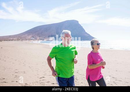 Ein paar von zwei reifen Menschen machen Bewegung und Sport zusammen Am Strand lächelnd und lachend - aktive Senioren tun Joggen und Laufen, um gesund und fit zu sein Stockfoto