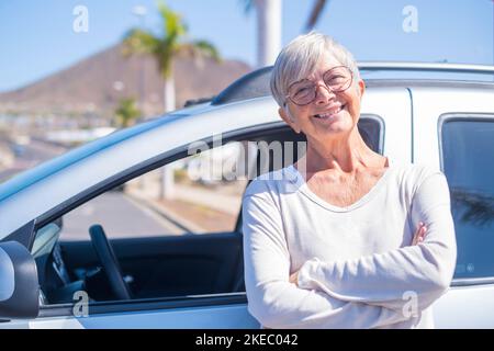 Nahaufnahme einer alten Frau, die lächelnd auf die Kamera blickt und ein Auto im Rücken hat. Porträt einer kaukasischen Frau, die neues Auto genießt. Zufriedener und zufriedener Kunde Stockfoto