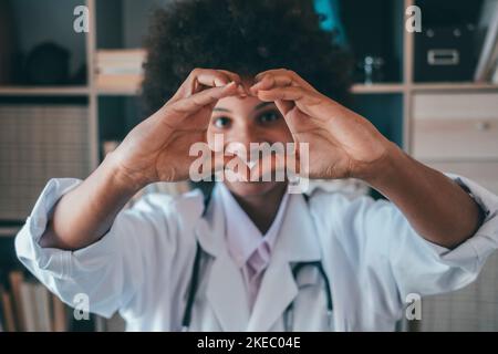 Frau tut Herzform Geste mit den Händen. Lächelnde schwarze Ärztin, die mit ihren Händen ein Liebessymbol macht. Mitarbeiter im Gesundheitswesen, der den Patienten Liebe und Unterstützung gibt Stockfoto