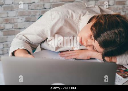Eine wunderschöne weibliche Teenager schläft und ruht sich auf dem Tisch, während sie zu Hause studiert oder arbeitet. Millenial macht eine Pause nach Hausaufgaben mit dem Laptop. Stockfoto