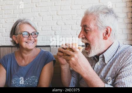 Ein paar Senioren zusammen essen und lachen - Reifer Mann hält einen Hamurger und isst ihn mit dem Mund Öffnete sich und seine Frau sah und lächelte Stockfoto