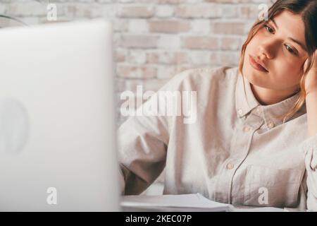 Junge Studentin sitzt am Schreibtisch voll mit Büchern Lehrbücher Blick auf Computer-Bildschirm zu unfertigen Hausaufgaben fühlt sich müde gelangweilt und unmotiviert, Mädchen mit Aufgaben, Arbeitsstau Überlauf Konzept geladen Stockfoto