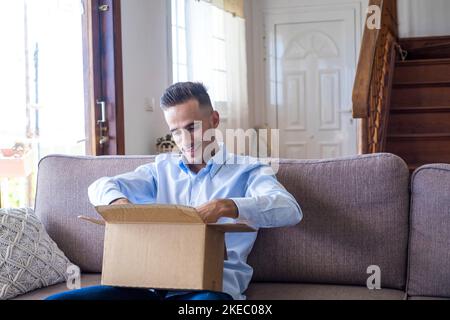 Mann Auspacken Delivdery Box öffnen Paket zu Hause. Glücklicher junger Mann, der zu Hause auf dem Sofa im Wohnzimmer auf den Karton schaute. Mann, der auscheckte, brachte Sachen zu Hause an. Stockfoto