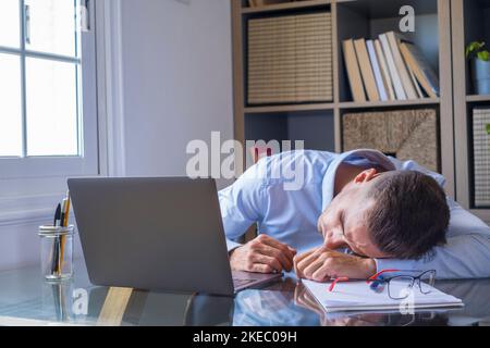 Der müde Geschäftsmann schläft an seinem Schreibtisch mit einem Laptop. Erschöpfter kaukasischer Kerl, der mit geschlossenen Augen auf dem Tisch lag und einschlief. Männliche Führungskraft schläft am Arbeitsplatz, langweilige Routinearbeit Stockfoto
