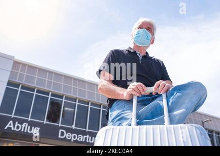 Ein reifer Mann trägt medizinische Maske mit seinem Gepäck oder Trolley Reisen und Flüge am Flughafen - Senior Warten auf seinen Flug verhindern Coronavirus oder eine andere Art von Virus (Covd-19) Stockfoto