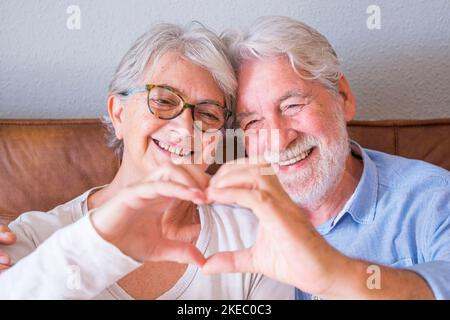 Porträt eines fröhlichen älteren Ehepaars, das sich umarmt und mit den Händen eine herzförmige Geste macht. Ältere glückliche Paar entspannen und posieren vor der Kamera sitzen im Wohnzimmer. Stockfoto