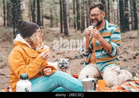Zwei glückliche Menschen und ein Hund essen zusammen und machen ein Picknick auf dem Boden in den Bergen mit vielen Bäumen um sie herum Stockfoto