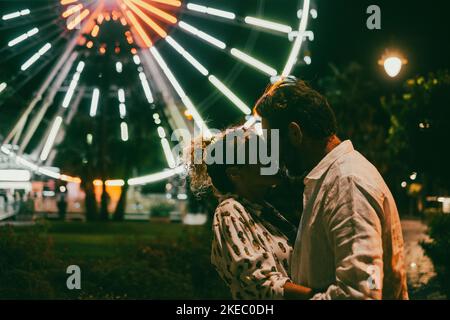 Liebevolles Paar umarmt sich leidenschaftlich auf der Straße gegen das beleuchtete Riesenrad in der Nacht. Paar romancing und verbringen gute Zeit im Freien während des Urlaubs in der Nacht Stockfoto