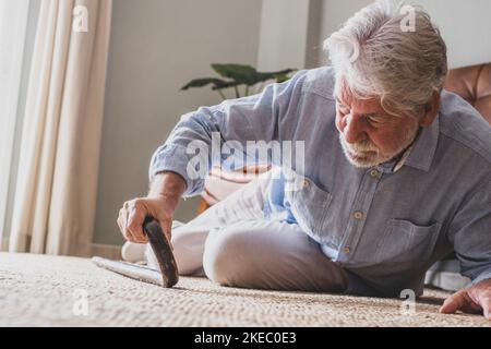 Älterer Mann, der auf dem Boden lag, nachdem er mit einem hölzernen Spazierstock neben der Couch auf dem Teppich im Wohnzimmer zu Hause heruntergefallen war. Alter Mann, der unter Schmerzen leidet und nach einem Sturz zu Hause um aufzustehen kämpft Stockfoto