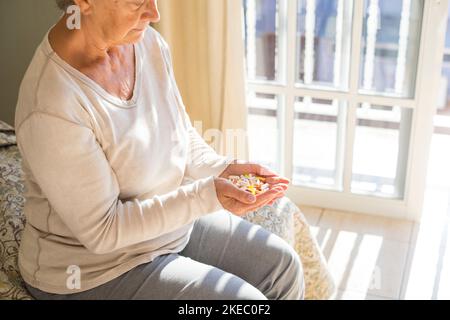 Ältere ältere Frau, die bunte Tabletten, Pillen und Kapseln in geschälten Händen ansieht, während sie zu Hause auf dem Bett sitzt, während sie nachdenkt, während sie Medizin einnimmt. Frau mit vielen Medikamenten in ihren Händen Stockfoto