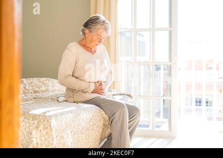 Ältere Frau, die am Morgen auf dem Bett zu Hause Bauch mit Bauchschmerzen hielt. Alte pensionierte Frau, die auf dem Bett saß und ihren Magen berührte, fühlte sich im Schlafzimmer zu Hause unwohl Stockfoto