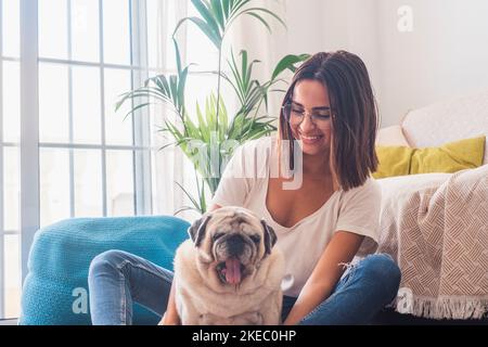 Porträt einer schönen Frau in einer Brille, die sich mit ihrem Hund, der auf dem Boden sitzt, im Wohnzimmer ihres Hauses amüsieren kann. Fröhliche Frau verbringt Freizeit mit ihrem niedlichen Haustier Hund zu Hause Stockfoto