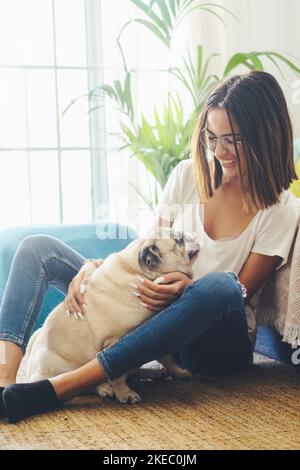 Porträt einer schönen Frau in einer Brille, die sich mit ihrem Hund, der auf dem Boden sitzt, im Wohnzimmer ihres Hauses amüsieren kann. Fröhliche Frau verbringt Freizeit mit ihrem niedlichen Haustier Hund zu Hause Stockfoto