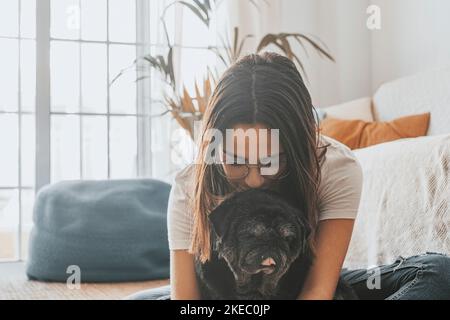 Porträt einer schönen Frau in einer Brille, die sich mit ihrem Hund, der auf dem Boden sitzt, im Wohnzimmer ihres Hauses amüsieren kann. Fröhliche Frau verbringt Freizeit mit ihrem niedlichen Haustier Hund zu Hause Stockfoto