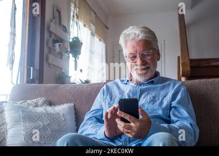 Der alte Mann sitzt lächelnd auf dem Sofa im Wohnzimmer und hält das Telefon, genießt es, mit dem Smartphone zufrieden zu sein, Nachrichten zu senden, Freunde anzurufen, im Internet zu surfen Stockfoto