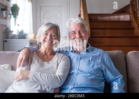 Porträt eines glücklichen Rentnerpaares mittleren Alters, das sich zu Hause auf dem gemütlichen Sofa entspannt. Lächelnd aufrichtig liebende ältere Hausbesitzer, die Kamera betrachten, für Fotos posieren, Liebe und Fürsorge in Innenräumen zeigen. Stockfoto