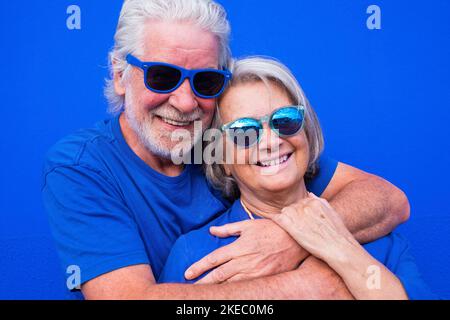 Portrait von ein paar reifen Menschen oder Senioren zusammen Spaß lächeln und die Kamera betrachten - Frau und Mann Rentner in blauem Hemd und Sonnenbrille mit blauem Hintergrund - trendige bunte und glückliche Menschen Stockfoto