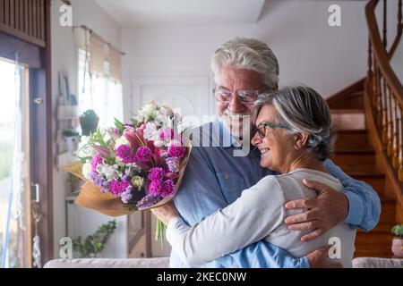 Porträt von zwei glücklichen und verliebten Senioren oder reifen und alten Menschen, die Blumen zu Hause halten und nach draußen schauen. Rentner Erwachsene genießen und feiern Urlaub zusammen. Stockfoto