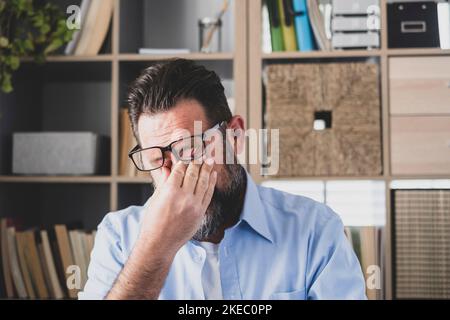 Ungesund gestresster Geschäftsmann, der Brillen auszieht, Augenlider reibt, an einem trockenen Augen-Syndrom leidet, das durch lange Computerüberarbeit verursacht wird, massiert Nasenrücken, um Schmerzen im Büro zu Hause zu lindern Stockfoto