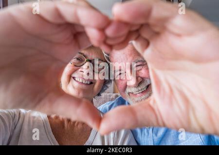 Nahaufnahme Porträt glücklich aufrichtig im mittleren Alter ältere Rentnerfamilie Paar macht Herzgeste mit den Fingern, zeigt Liebe oder zeigt aufrichtige Gefühle zusammen drinnen, Blick auf die Kamera. Stockfoto