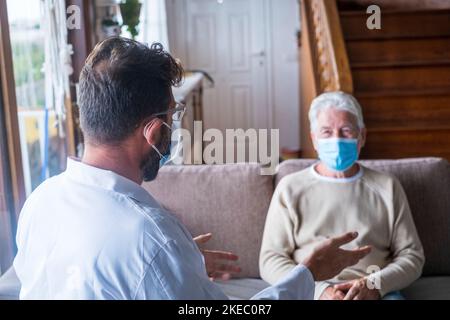 Männliche professionelle Arzt Beratung Senior Patient während der medizinischen Versorgung Besuch trägt Masken. Ein junger Arzt und ein alter älterer Herr, der auf dem Sofa sitzt und medizinische Hilfe leistet. Ältere Menschen in der häuslichen Pflege. Covid- und Coronavirus-positiver Test Stockfoto