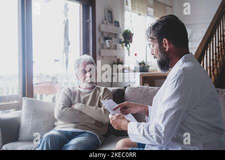 Männlicher professioneller Arzt, der während eines Arztbesuchs einen älteren Patienten konsultiert. Ein junger Arzt und ein alter älterer Herr, der auf dem Sofa sitzt und medizinische Hilfe leistet. Konzept für die häusliche Pflege älterer Menschen Stockfoto