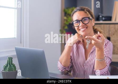 Glückliche Frau, die die Schlüssel ihres neuen Hauses oder Büros zeigt. Porträt einer lächelnden Geschäftsfrau mit Schlüssel und Laptop auf dem Schreibtisch. Mieterin oder Mieterin, die mit Schlüsseln in der Hand umzieht oder umzieht Stockfoto