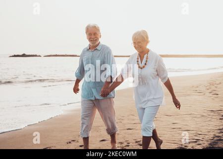 Ein paar ältere Leute, die gemeinsam auf dem Sand spazieren und sich am Strand amüsieren und den Moment genießen. Zwei niedliche Senioren in der Liebe Spaß haben. Barfuß auf dem Wasser laufen Stockfoto