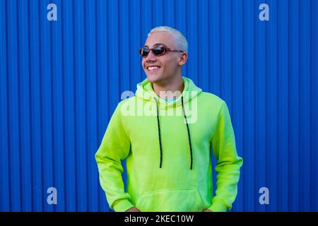 Porträt eines jungen Mannes oder Teenagers, der lächelt und Spaß hat, indem er trendige Sonnenbrillen und einen Limettenpullover mit blauem, farbenfrohem Hintergrund trägt - fröhliche Menschen, die tausendjährig Freude haben Stockfoto