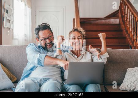 Erstaunte kaukasische Paar feiert Online-Erfolg mit Laptop sitzen auf dem Sofa zu Hause. Fröhliche Frau und Mann überrascht durch gute unglaubliche Nachrichten, riesige Shopping-Angebot auf der Website Stockfoto