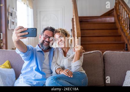 Glückliches kaukasisches Paar, das zu Hause mit der Handykamera auf dem Sofa sitzt und Selfie macht. Fröhliche Ehefrau und Ehemann, die sich auf dem Smartphone porträtieren. Pärchen genießen das Klicken auf Bilder in der neuen Wohnung Stockfoto
