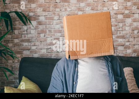 Reifer Mann in leerem Karton sitzt auf dem Sofa im Wohnzimmer des Hauses. Kaukasischer Kerl mit Gesicht versteckt oder in Karton-Lieferbehälter bedeckt, während Sie sich auf der Couch zu Hause entspannen Stockfoto