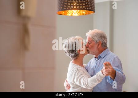 Ein romantisches, liebevolles Seniorenpaar, das sich beim Küssen die Hände hält und gemeinsam im Wohnzimmer des Hauses tanzt, ein älteres, glückliches Paar, das zu Hause tanzt. Alter Mann und Frau haben Spaß Zeit in der modernen Wohnung Stockfoto