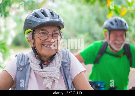 Porträt einer alten Frau, die lächelt und die Natur im Freien genießt, mit ihrem Mann, der lachend Fahrrad fährt. Kopfschuss einer reifen Frau mit Brille, die sich gesund anfühlt. Stockfoto