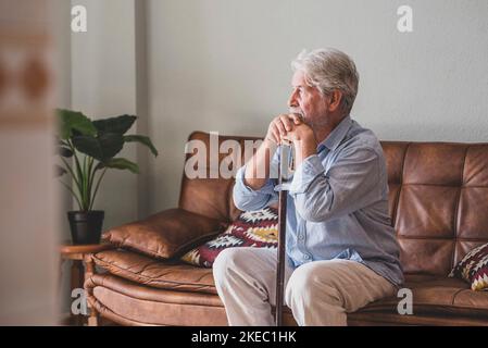 Nachdenklicher älterer Mann, der sich auf dem Sofa auf einen Spazierstock lehnte. Einsamer älterer Mann, der zu Hause im Wohnzimmer sitzt. Ein alter Mann mit grauen Haaren, der sich auf einen Spazierstock lehnte und zu Hause auf der Couch saß Stockfoto