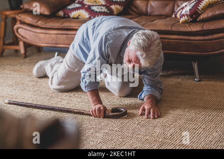 Älterer Mann, der auf dem Boden lag, nachdem er mit einem hölzernen Spazierstock neben der Couch auf dem Teppich im Wohnzimmer zu Hause heruntergefallen war. Alter Mann, der unter Schmerzen leidet und nach einem Sturz zu Hause um aufzustehen kämpft Stockfoto