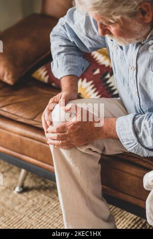 Älterer alter Mann, der mit Knieproblemen konfrontiert ist und zu Hause auf dem Sofa sitzt und das Knie hält. Alter Mann, der an schweren Gelenkschmerzen leidet, sitzt im Wohnzimmer. Gesundheitswesen und Problemkonzept Stockfoto