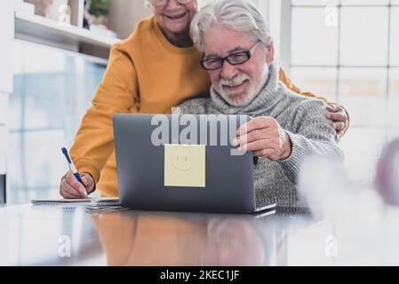 Ein paar von zwei glücklichen Senioren, die zu Hause zusammen arbeiten und mit einem Laptop arbeiten, lächeln und Spaß zusammen haben - Lockdown Lifestyle Stockfoto