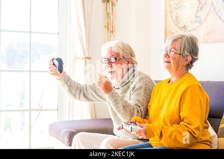 Ein paar Senioren und ältere Menschen, die auf dem Sofa und dem fernseher schauten und die Controller benutzten, die Videospiele zu Hause zusammen spielten - ein alter Mann, der beim Spiel gewann Stockfoto