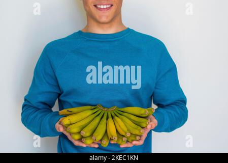 Porträt und Nahaufnahme von Mann oder Teenager oder jung Junge hält eine Menge Bananen und lächelt und schaut zu Die Kamera - Obst halten - gesunde Lebensweise und Konzept Und Eigenschaften der Banane Stockfoto