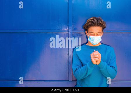 Porträt und Nahaufnahme eines Teenagers, der betet und eine medizinische und chirurgische Maske auf seinem Gesicht trägt - tausendjähriges Gebet mit geschlossenen Augen, blauem Pullover und blauem Hintergrund Stockfoto