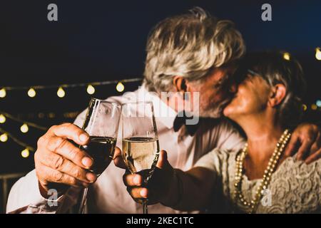 Altes kaukasisches Paar küsst sich beim Toasten von Sektgläsern zur Feier der Neujahr-Party. Liebevoller Ehemann und Ehefrau küssen sich beim Feiern mit Weißweingläsern in den Händen Stockfoto