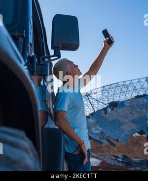 Ein junger Mann oder Teenager, der mit seinem Telefon und seinem Auto nach einem Signal sucht - tausendjährig süchtig am Telefon im Nirgendwo auf der Suche nach einer Verbindung - blondes Haar erwachsen Stockfoto