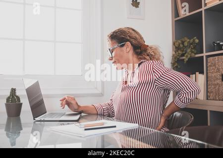 Kaukasische Frau, die ihren Rücken wegen Stressschmerzen und Krankheiten berührt, die durch die Arbeitshaltung verursacht werden. Eine Geschäftsfrau leidet unter Rückenschmerzen, während sie auf einem Bürostuhl sitzt und am Laptop arbeitet. Weibliche Führungskraft mit Schmerzen im unteren Rücken Stockfoto