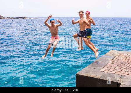 Eine Gruppe von Freunden, die zusammen am Strand abspringen, Flips machen und Spaß im Wasser haben - die Leute genießen ihren Urlaub am Strand beim Spielen und Lachen - beim Springen auf die Kamera schauen Stockfoto