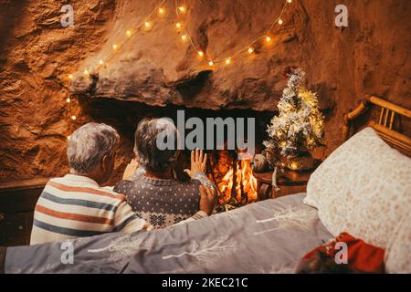 Altes kaukasisches Paar, das die Hände zu Hause warm hält. Ehemann und Ehefrau im älteren Alter entspannen sich, während sie die Handfläche zum Erwärmen am brennenden Kamin während der winterweihnachtsferien öffnen Stockfoto